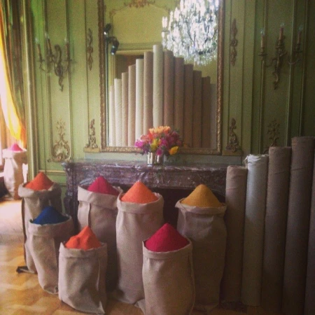 Bags of colorful pigment or dye line a room with decorative molding and a chandelier, rolls of carpet stand against the back wall.