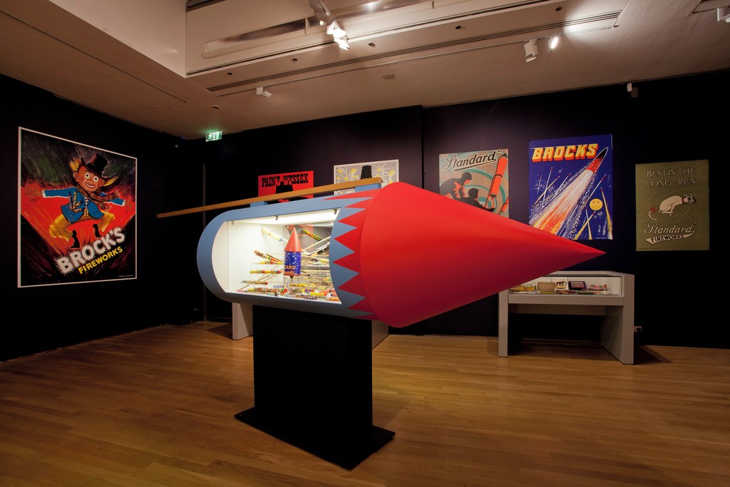 Interior shot of an exhibition space featuring a rocket-shaped display case filled with colorful fireworks, with vintage posters on the walls.
