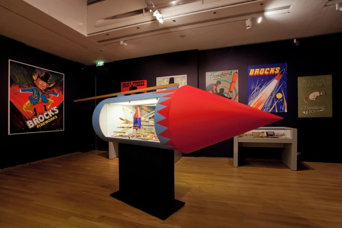 Interior shot of an exhibition space featuring a rocket-shaped display case filled with colorful fireworks, with vintage posters on the walls.