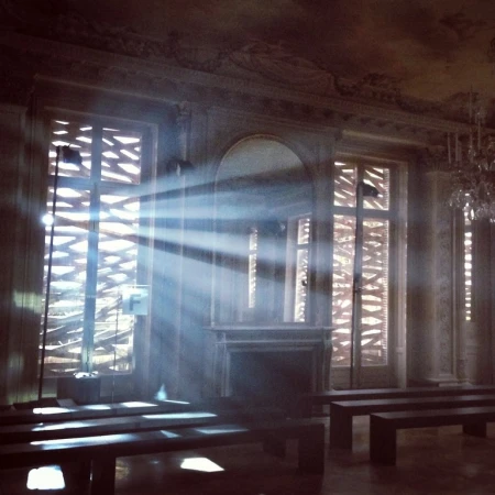 Ornate room interior with sunlight streaming through lattice windows, casting shadows on benches and highlighting architectural details.