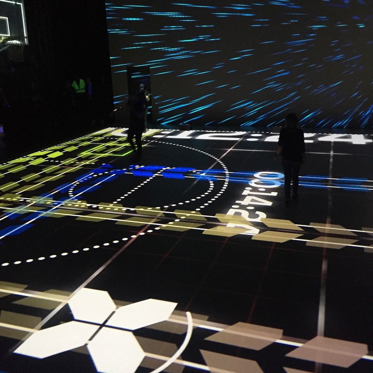 An indoor interactive exhibit features graphics projected onto the floor with two adults standing on the projected design, creating a dynamic, modern display.
