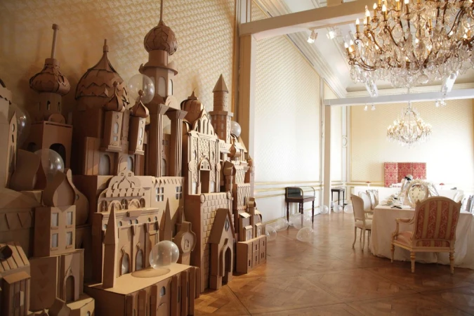 Intricate cardboard castle art installation with clear balloons inside formal dining room with chandeliers, table settings and parquet floor.