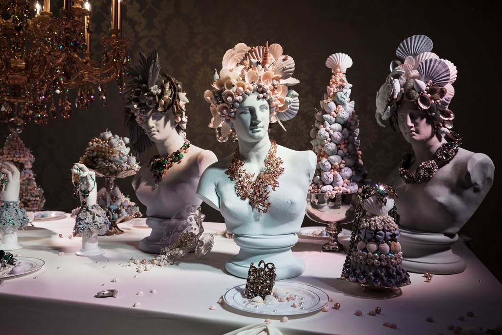 Bust sculptures wearing seashell headpieces and necklaces on a table under a chandelier, showcasing unique art and design in an indoor setting.