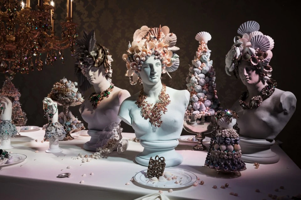 Bust sculptures wearing seashell headpieces and necklaces on a table under a chandelier, showcasing unique art and design in an indoor setting.