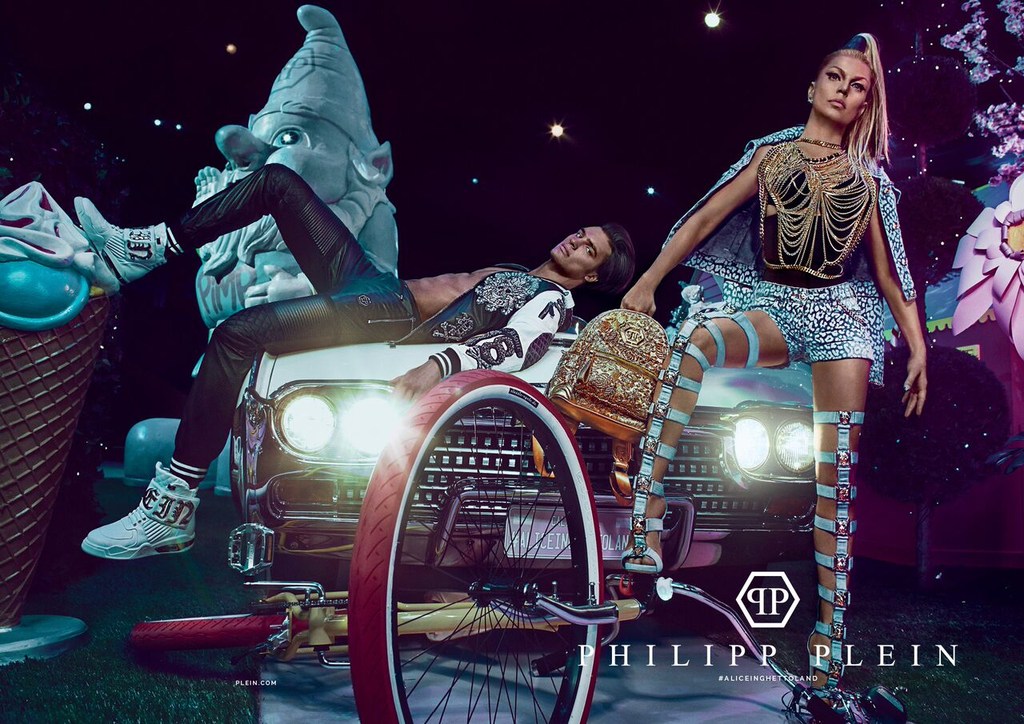 Fashion shot in whimsical setting features two models, one lying on a car hood, the other standing, accessorized with a backpack, in night.