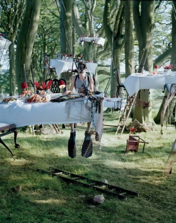 Woman in snorkeling mask sits among tables with seafood and other items in a surreal forest picnic scene, editorial photography.