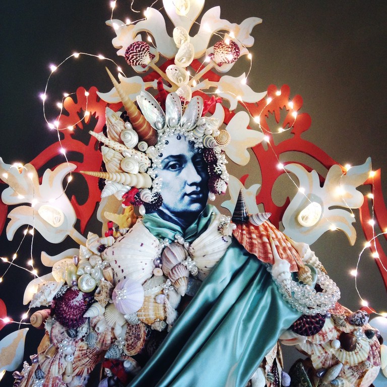 Portrait of a figure adorned with seashells and pearls, creating a whimsical and ornate costume against a dark background with fairy lights.