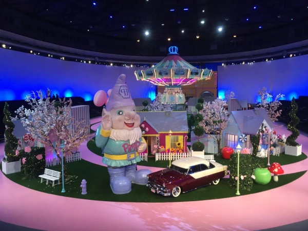 Whimsical scene with gnome statue, vintage car, and carousel against a blue backdrop. Playful, fantasy-themed arrangement.