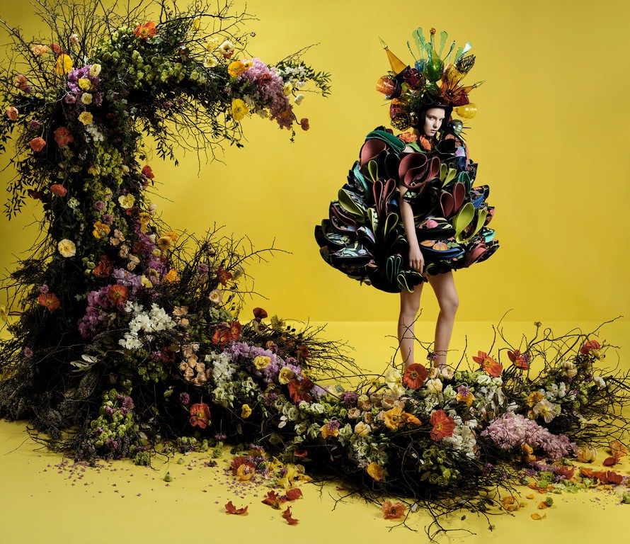 A woman in a floral dress poses in front of a yellow backdrop with a crescent-shaped floral arrangement, fashion photography.