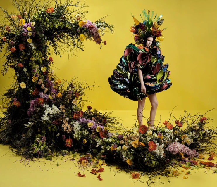 A woman in a floral dress poses in front of a yellow backdrop with a crescent-shaped floral arrangement, fashion photography.