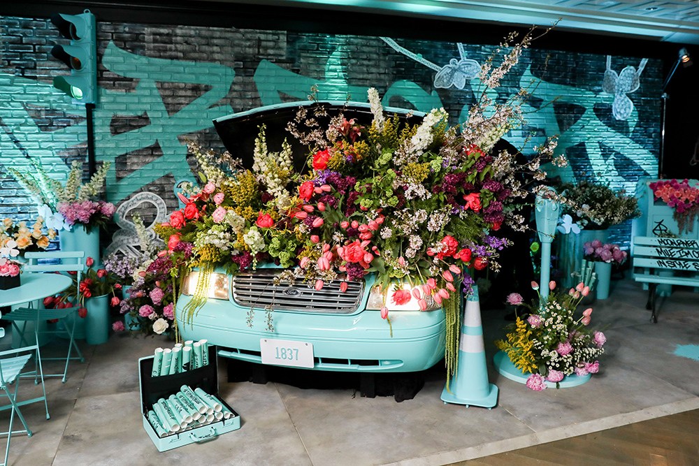 Car covered in vibrant flowers at event, with traffic light prop and sign, creating a whimsical and artistic display.