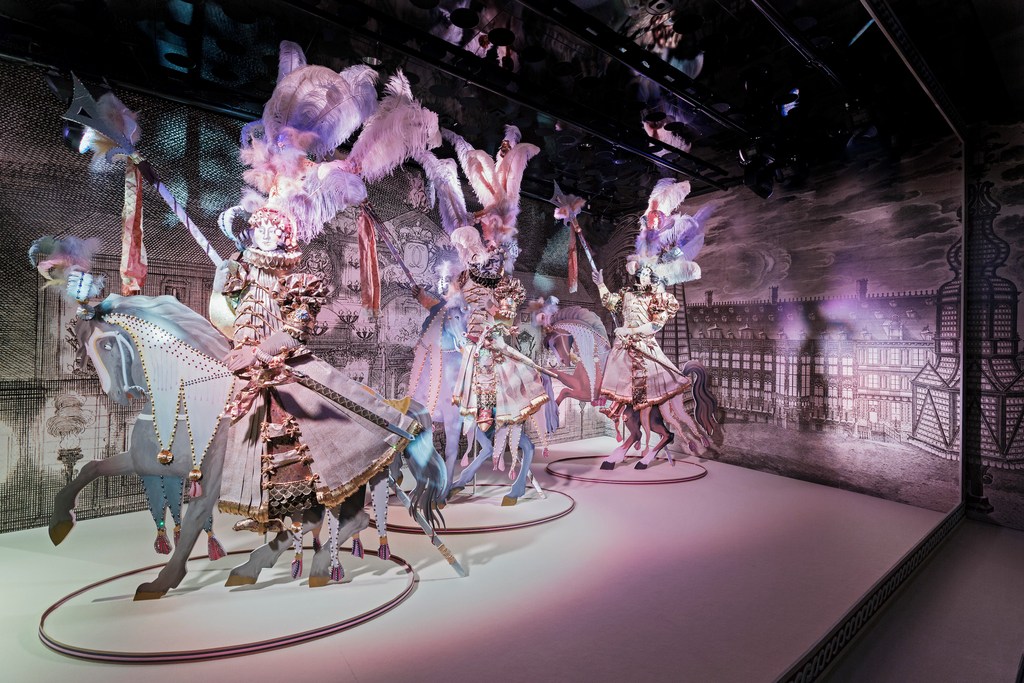 Figures of people riding horses are displayed on a stage in an indoor setting with an ornate background design; theatrical art exhibit.