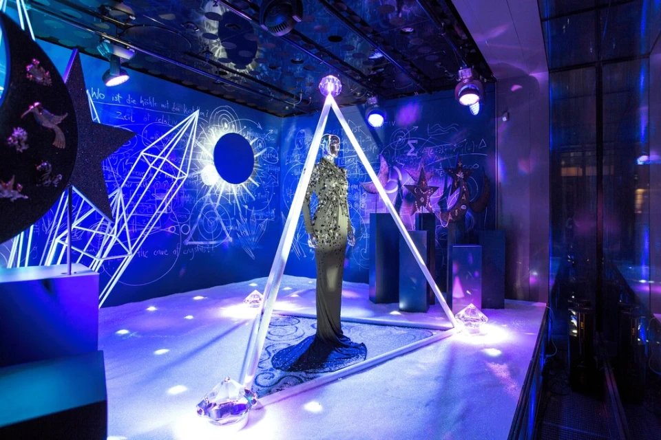 Mannequin in an ornate dress inside a triangular frame in a room with stars, moons, and geometric designs on walls and ceiling.