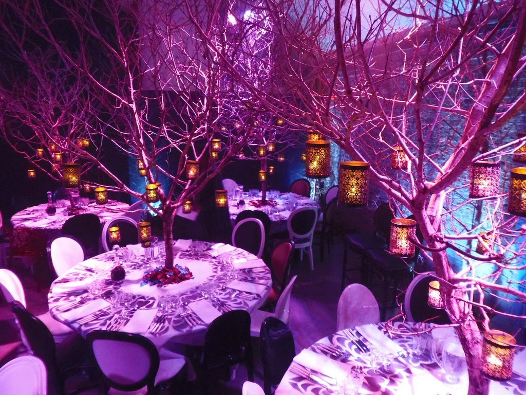 Interior scene with decorated tables, bare trees with hanging lanterns, and ambient pink-purple lighting creates a whimsical atmosphere.