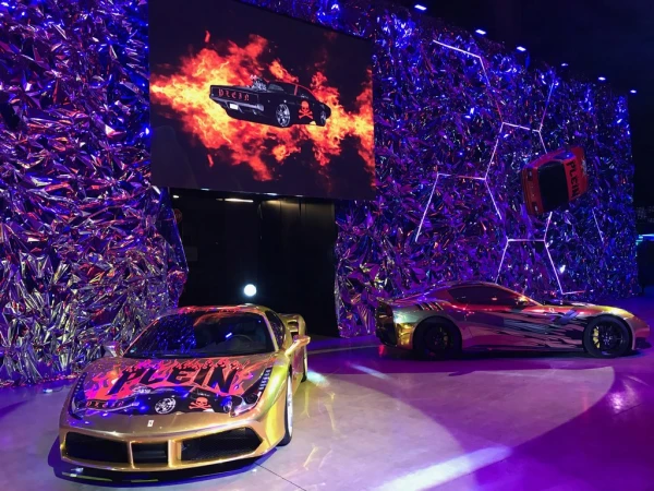 Two modified luxury sports cars showcased on a stage with a shiny textured backdrop, bathed in blue and purple lighting, evoking a modern automotive event.