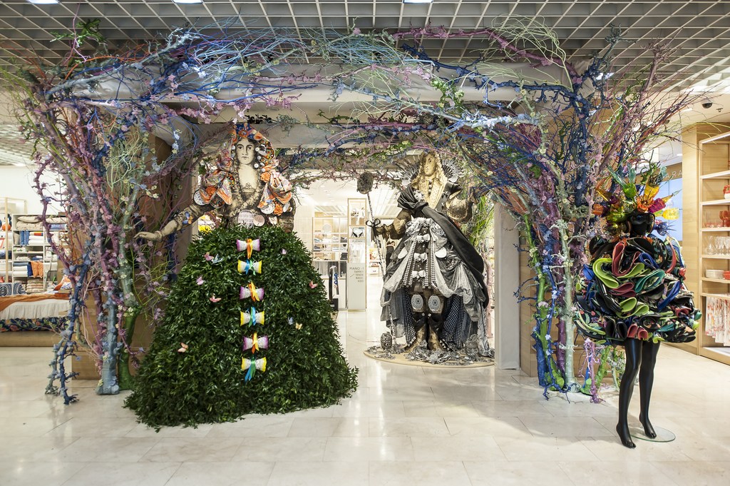 Store window display features mannequins in artistic foliage and fabric creations, framed by vibrant, painted tree branch structures.
