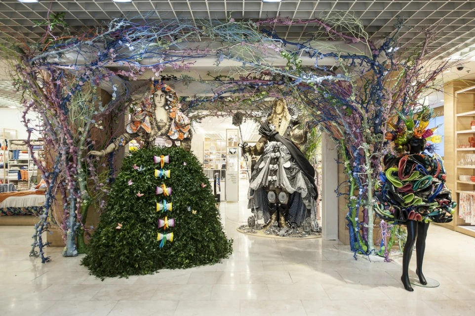 Store window display features mannequins in artistic foliage and fabric creations, framed by vibrant, painted tree branch structures.