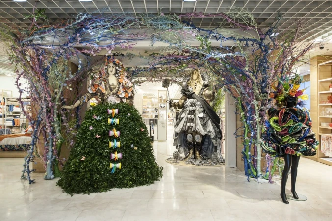 Store window display features mannequins in artistic foliage and fabric creations, framed by vibrant, painted tree branch structures.