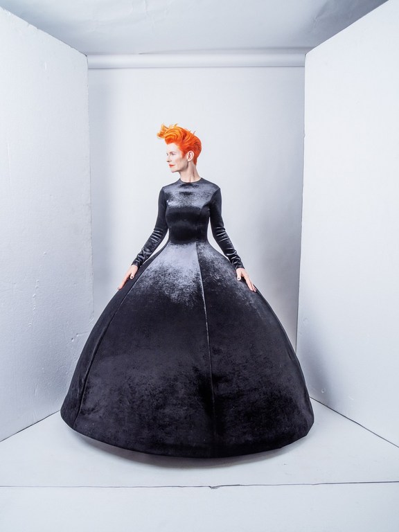 Woman with orange hair poses in a long-sleeved black velvet gown with a full skirt in a white corner photo studio setting.