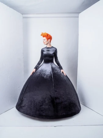 Woman with orange hair poses in a long-sleeved black velvet gown with a full skirt in a white corner photo studio setting.