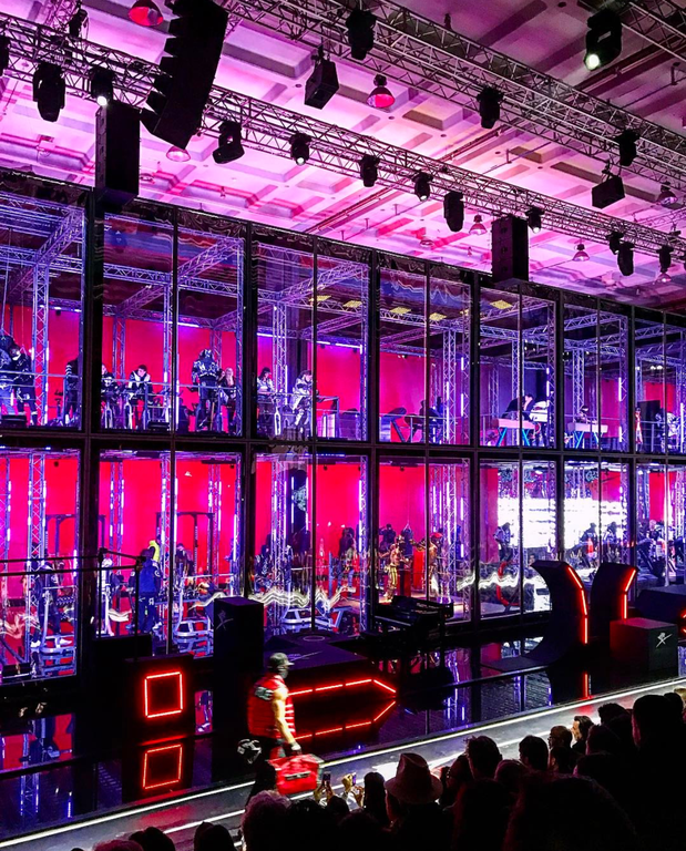 Wide shot of a musical performance with a large group of people performing inside individual glass enclosures on a stage with audience.