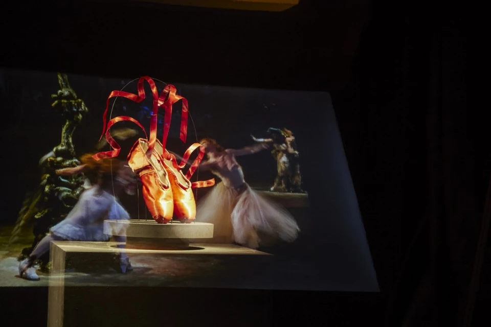 Ballet pointe shoes with red ribbons rest on a pedestal, dancers and a statue blurred in the background, against dark lighting.