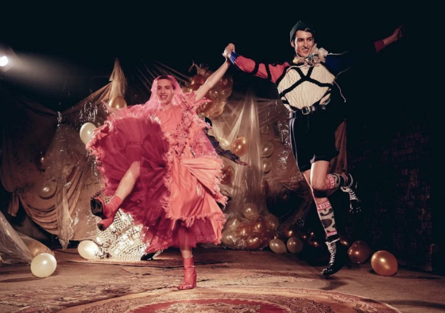 Two figures in artistic costumes dance in a spotlight on stage with balloons and draped plastic sheeting in the background.