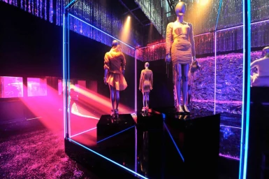 Mannequins on black pedestals inside a glowing neon frame under pink and blue stage lighting