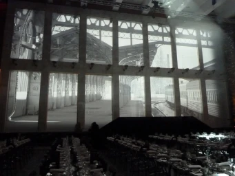 Large-scale black and white video projection of a railway station on a high stone wall above candlelit dining tables