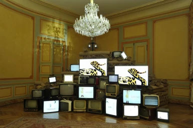 An elaborate stack of vintage televisions and monitors displaying leopard print graphics inside an ornate yellow room with a crystal chandelier.