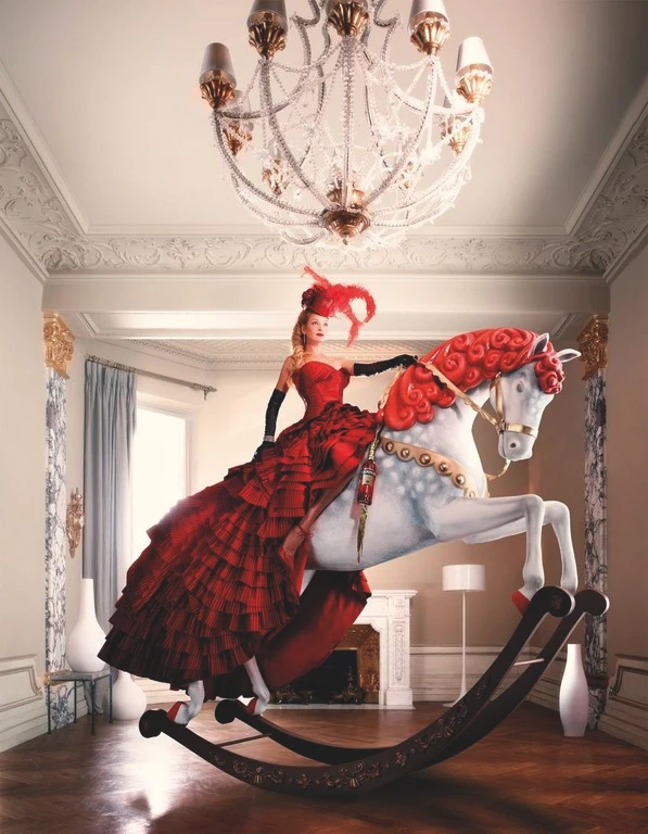 Woman in a red gown and feathered hat riding a large decorative white rocking horse beneath a grand crystal chandelier.