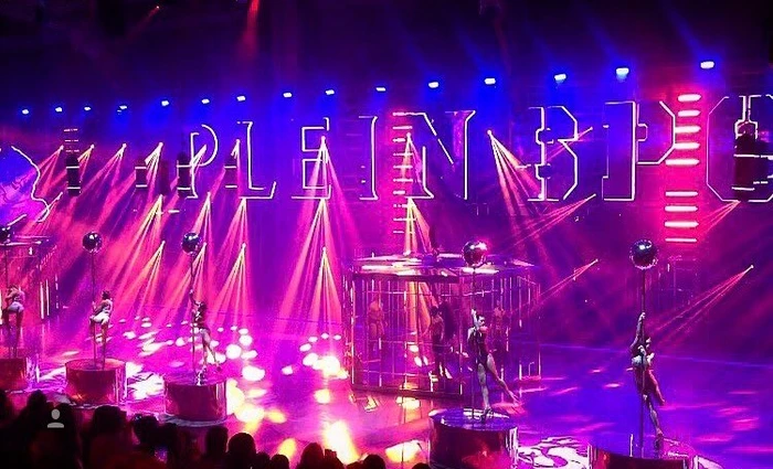 Stage performance with dancers on multiple vertical poles and a central cage under vibrant pink and blue lighting designed by Simon Costin
