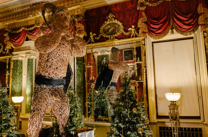 Large woven figure made of twigs with horns displayed in an ornate room with red damask walls and holiday trees by Simon Costin
