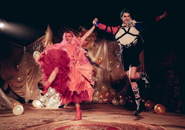 Two performers dancing and holding hands in a rustic space decorated with balloons and draped fabric, art directed by Simon Costin.