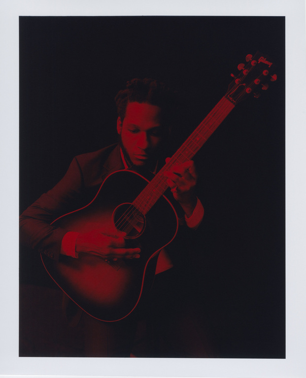 Musician in suit playing acoustic guitar with red lighting against a black background in an indoor setting in a portrait photograph.