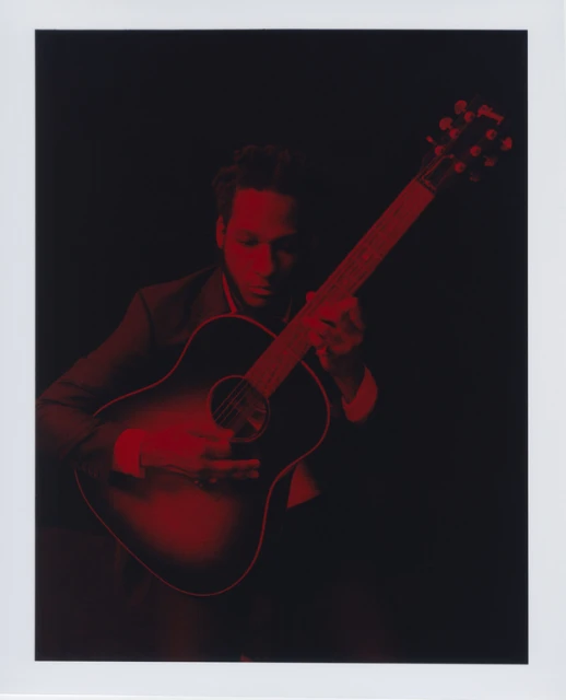 Musician in suit playing acoustic guitar with red lighting against a black background in an indoor setting in a portrait photograph.