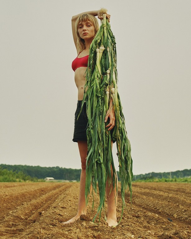 Adult woman in a red top and black shorts, standing barefoot in a brown field holding a large green onion, cloudy sky, nature, summer.