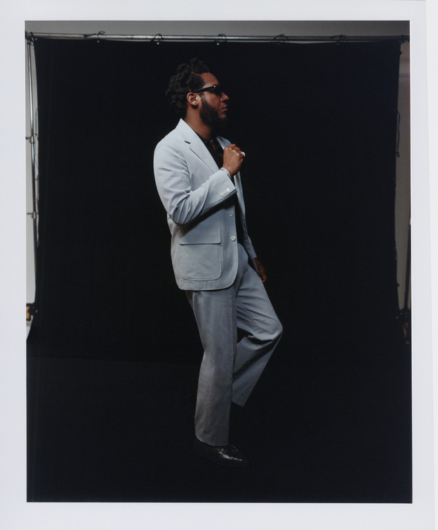 An adult male poses in a light grey suit against a black background. He's captured in profile, wearing sunglasses and black shoes.