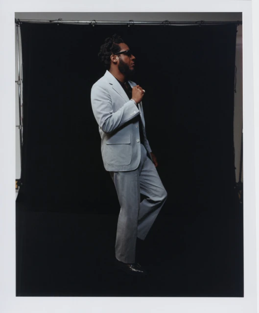An adult male poses in a light grey suit against a black background. He's captured in profile, wearing sunglasses and black shoes.