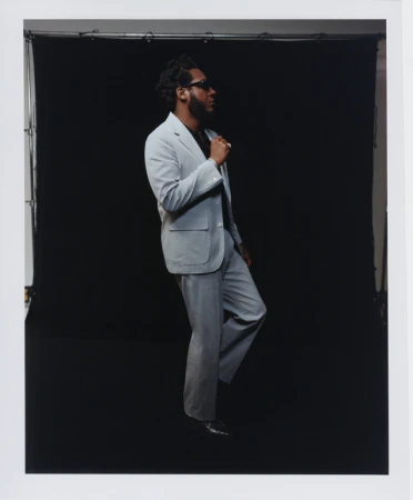 An adult male poses in a light grey suit against a black background. He's captured in profile, wearing sunglasses and black shoes.