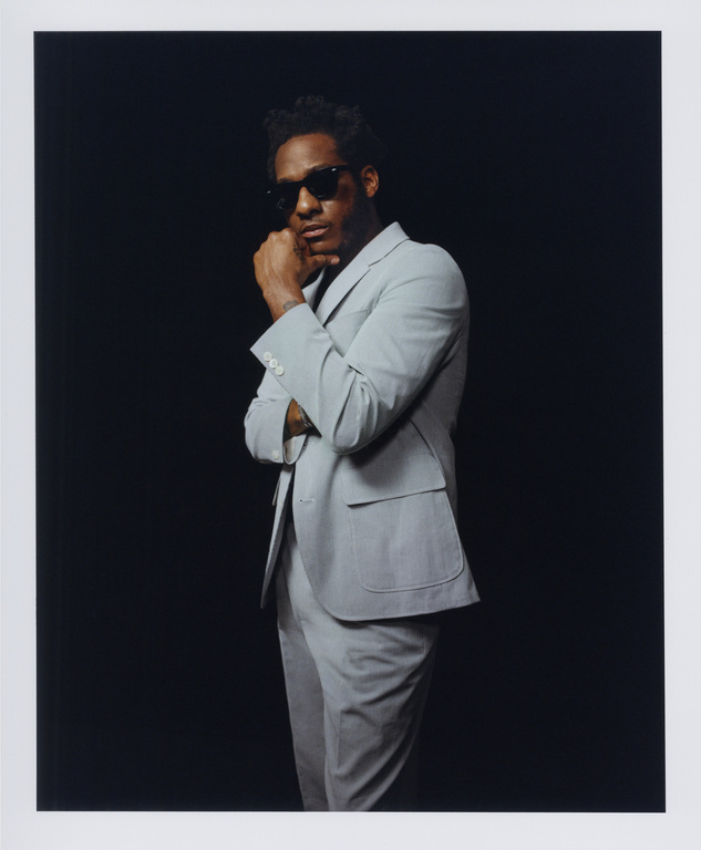 Man wearing a light grey suit and sunglasses posing against a dark background with a white border framing the image.