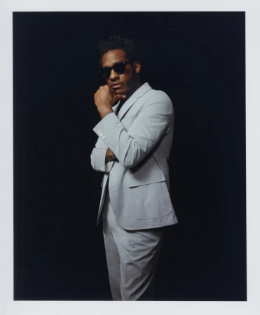 Man wearing a light grey suit and sunglasses posing against a dark background with a white border framing the image.