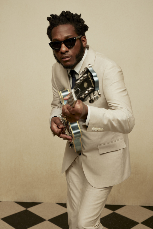 Stylish African-American musician in a tan suit and sunglasses playing an electric guitar in an indoor setting with a checkered floor.