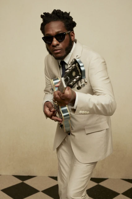 Stylish African-American musician in a tan suit and sunglasses playing an electric guitar in an indoor setting with a checkered floor.