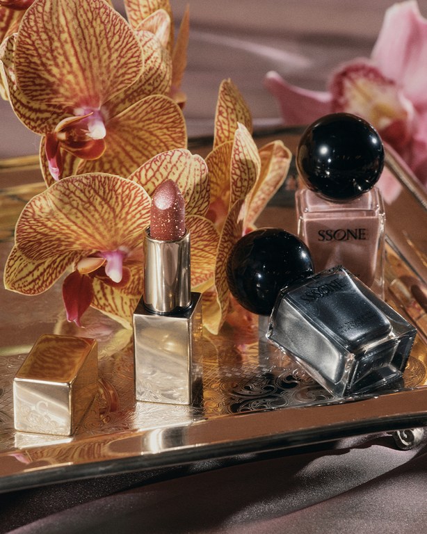 Still life shot of lipstick, nail polish bottles, and orchid flowers on a silver tray, highlighting beauty and elegance in a studio.