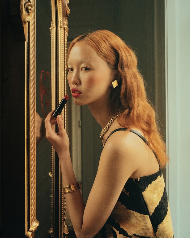 Asian woman applies red lipstick, reflected in a gilded mirror. Gold jewelry and black-and-gold dress. Indoor setting.
