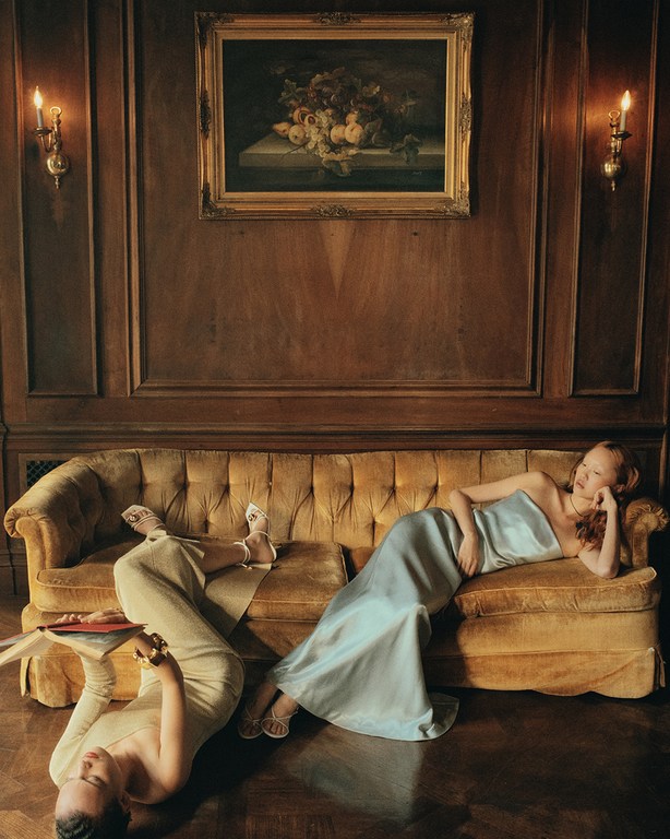 Two women lounge on a vintage gold couch in an ornate, wood-paneled room with a painting and sconces. One woman reads a book.