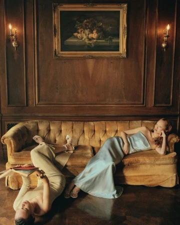 Two women lounge on a vintage gold couch in an ornate, wood-paneled room with a painting and sconces. One woman reads a book.