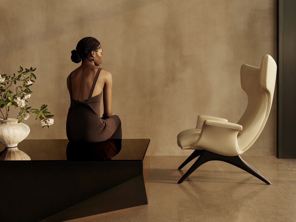Woman sits on a dark table next to an off-white high back chair. A vase of flowering branches sits on the table.
