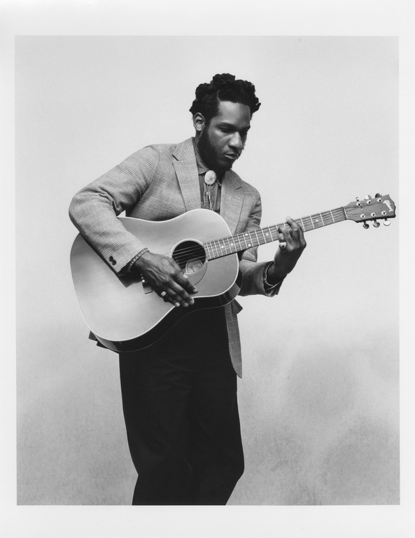 Black and white portrait of African American male playing an acoustic guitar, wearing a blazer and bolo tie, against a neutral background.